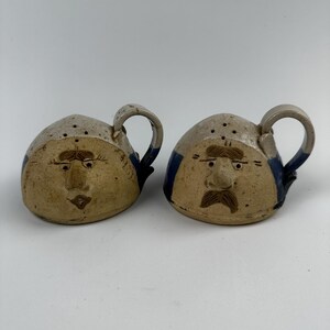May include: A set of two ceramic salt and pepper shakers with whimsical faces. The shakers are hand-painted with brown and blue details and have a rustic, handmade look.