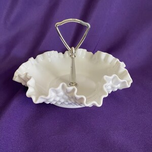 May include: A white, ruffled-edge serving dish with a silver handle. The dish has a textured, hobnail-like pattern on the base. The handle is a simple, arched design. The dish is set against a purple background.