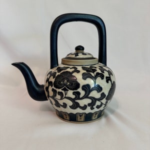 May include: A white ceramic teapot with a black floral design and a black handle. The teapot has a lid with a knob on top.