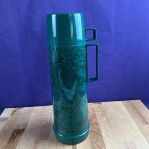 May include: A teal-colored thermos with a handle and a map design. The thermos has a cylindrical shape with a base and a lid. The map design features a compass rose and nautical elements. The thermos is on a wooden surface.