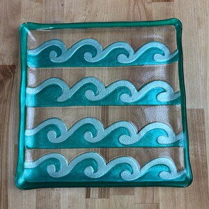 May include: Square glass serving dish with a teal green border and wave design. The waves are teal and silver, set against a light brown background. The dish is approximately 8 inches square.