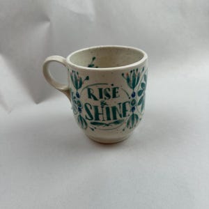 May include: A ceramic mug with a cream-colored speckled finish. The mug features a teal floral design and the words "RISE & SHINE" in teal script. The mug has a small handle and is suitable for hot or cold beverages.