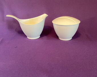 May include: A white ceramic creamer and sugar bowl set. The creamer is shaped like a bird with a curved spout. The sugar bowl has a rounded shape with a lid.