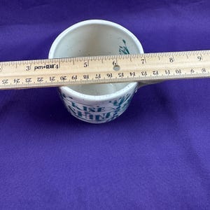 May include: A white ceramic mug with a speckled finish and a teal floral design. The mug has the words "RISE & SHINE" printed on the side. The mug is approximately 4 inches in diameter.