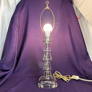 May include: A clear glass table lamp with a stacked geometric design. The lamp has a gold-colored harp and a round light bulb. The base and stacked elements are faceted, and the lamp is plugged into a wall outlet.