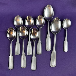May include: A collection of nine silver spoons arranged on a purple surface. The spoons vary in size, with oval bowls and long, slender handles. The handles have a decorative beaded border.