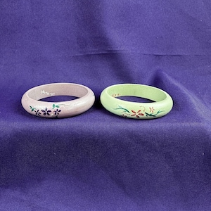 May include: Two wooden bangles. One is light pink with purple floral designs and the other is light green with red and green floral designs. The bangles are on a purple background.
