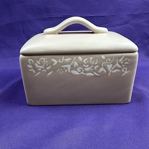 May include: A rectangular, beige ceramic container with a matching lid and handle. The front of the container features a white floral design. The lid has a curved handle for easy lifting. The container is likely for storing food or other small items.