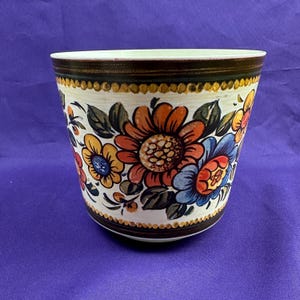 May include: A ceramic flower pot with a cream-colored base and a dark brown rim. The pot is decorated with colorful floral designs in orange, yellow, and blue. The flowers are surrounded by green leaves, creating a vibrant and eye-catching pattern.