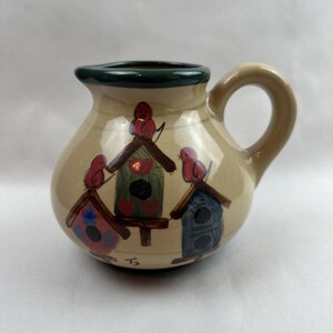 May include: A beige ceramic pitcher with a dark green rim and a curved handle. The pitcher is decorated with three birdhouses in red, green, and blue, each with a red bird perched on top. The base of the pitcher has the initials "T3".