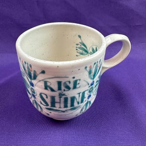 May include: A white ceramic mug with a speckled finish. The mug features teal floral designs and the words "RISE SHINE" in a stylized font. The mug has a curved handle and is set against a purple background.