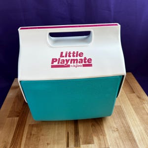 May include: A teal and white Little Playmate cooler by Igloo. The cooler has a white handle and a magenta logo. The cooler is sitting on a wooden surface.
