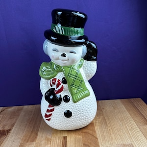 May include: A ceramic snowman figurine with a black top hat, green scarf, and black gloves. The snowman has a smiling face, black button eyes, and a candy cane. A festive holiday decoration.