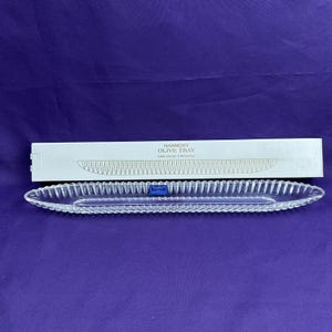 May include: A clear, elongated 24% lead crystal olive tray with a scalloped edge design. The tray is positioned in front of its original white rectangular box, which features the text Harmony Olive Tray 24% Lead Crystal and a line drawing.