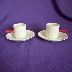 May include: Two white ceramic coffee cups with red handles and matching saucers. The cups and saucers have a thin gold rim.