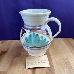 May include: Cream-colored ceramic pitcher with a wide handle and spout. The pitcher is decorated with blue, red, and green floral designs. A tag from Jamestown Pottery is visible.