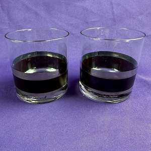 May include: Two clear glass tumblers with a dark band and silver trim. The tumblers have a cylindrical shape and the text "Georges Briard" is printed on the bottom. The glasses are on a purple surface.