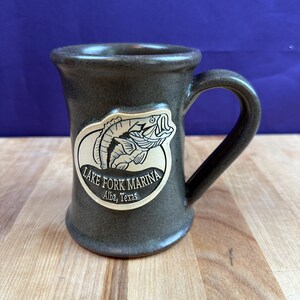 May include: A dark gray ceramic mug with a curved handle. The mug features a white and tan sticker with a fish illustration and the text "Lake Fork Marina Alba, Texas."