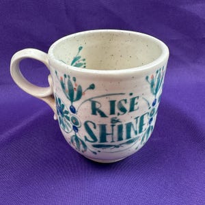 May include: A ceramic mug with a white speckled finish and a curved handle. The mug is decorated with teal and blue floral designs and the words "RISE & SHINE" in teal script. The mug is a standard size for coffee or tea.
