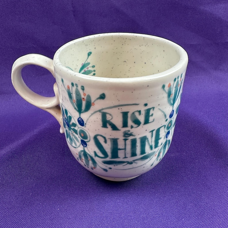 May include: A ceramic mug with a white speckled finish and a curved handle. The mug is decorated with teal and blue floral designs and the words "RISE & SHINE" in teal script. The mug is a standard size for coffee or tea.