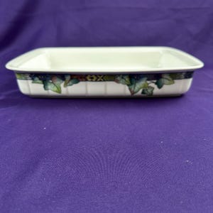 May include: Rectangular white ceramic baking dish with a decorative border featuring green ivy leaves and a geometric pattern. The dish has a slightly raised rim and is set against a purple background. A kitchenware item.