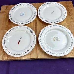 May include: Four white ceramic plates with cocktail-themed designs. Each plate features a different cocktail name and a related illustration: a sailboat, a nail, a screwdriver, and a cityscape. The text is printed in a black, stylized font.