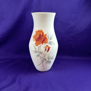 May include: A white vase with a gold rim and a painted orange rose design. The vase has a slender, curved shape and a wide opening. The rose is detailed with green leaves and a red bud.