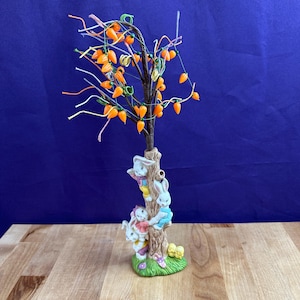 May include: A whimsical Easter figurine featuring a tree adorned with orange carrot ornaments and colorful string lights. Three bunny figures climb the tree trunk, set on a green base with a yellow bow. The background is a deep blue.