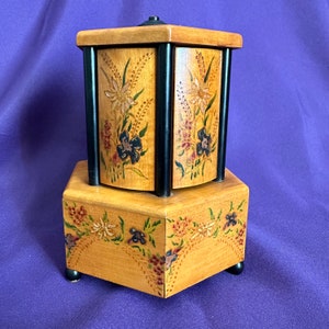 May include: A wooden hexagonal music box with a rotating top. The top is painted with floral designs in shades of yellow, white, and black. The base of the music box is also painted with floral designs in shades of red, white, and black.