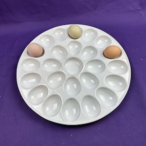 May include: A white ceramic egg holder with 20 egg-shaped indentations. Three eggs of varying colors are placed in the holder: light brown, light green, and brown. The egg holder is round and sits on a purple surface.