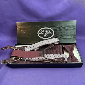 May include: A set containing a pair of burgundy and white patterned suspenders with dark brown leather tabs and a matching burgundy textured bow tie, presented in a black box labeled La Valière Paris on the lid interior.