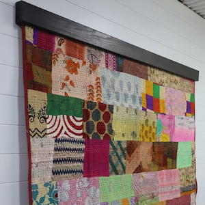 May include: A patchwork textile wall hanging with a dark wood frame. The textile is composed of various fabric squares in shades of red, pink, green, blue, and yellow. The design includes floral and geometric patterns. The wall hanging is mounted on a white wall.