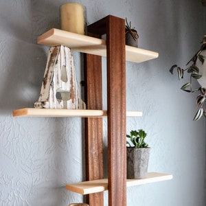 May include: A wooden shelf unit with five shelves. The shelves are made of light wood and the frame is made of dark wood. The shelves are decorated with a white wooden letter A, a potted succulent, a candle, and a wooden vase.