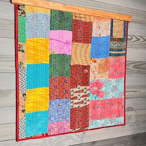 May include: A rectangular wall hanging featuring a patchwork of multi-colored fabric squares with various patterns including floral, geometric, and abstract designs. The textile is suspended from a light-toned wooden bar and has a red fabric border.