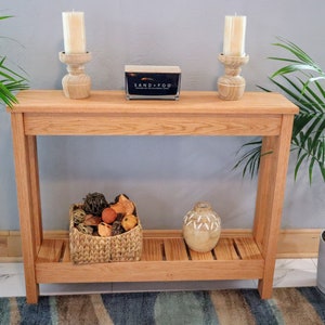 May include: A light-colored wooden console table with a lower shelf, holding a woven basket and a decorative vase. Two wooden candle holders with white candles and a small box with the text "SAND+FOG" are on the top surface. Green plants flank the table.