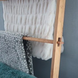 May include: A wooden ladder style blanket rack with two white blankets and a gray patterned blanket draped over the top rung.