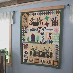 May include: A colorful quilt with a green border hangs on a wall. The quilt is made up of different squares with various designs, including a house, flowers, a sheep, and the words "Love Blooms Here".