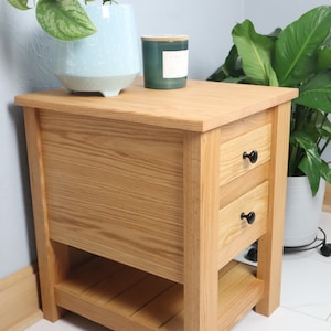 May include: A light wood end table with two drawers and a lower shelf. The table has black knobs on the drawers. A potted plant and a candle are on top of the table.