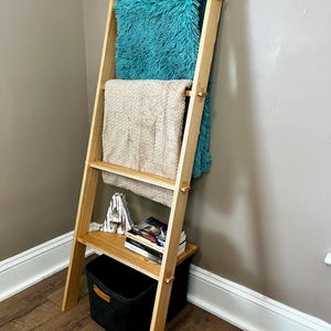 May include: A wooden leaning ladder shelf with a light finish. It holds a teal fluffy throw blanket, a beige knitted blanket, books, and a black storage basket. The shelf is leaning against a neutral-colored wall.