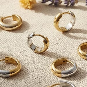 May include: A collection of six small hoop earrings, each featuring a two-tone design with one half in a polished gold finish and the other half in a silver finish. The earrings are circular with a hinged closure mechanism visible on each piece.