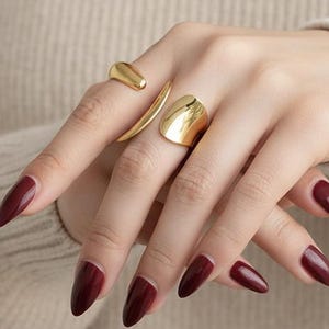 Peut inclure: Une main ornée de trois bagues dorées aux designs variés. Une bague présente une bande incurvée, une autre une forme arrondie et la troisième un design lisse et ouvert. Les ongles sont peints d'un bordeaux profond, contrastant avec les bijoux dorés.