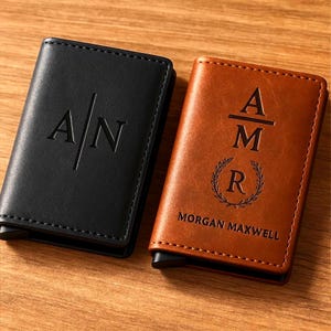 Könnte beinhalten: Zwei personalisierte Ledergeldbörsen. Eine ist schwarz mit den eingravierten Initialen "A/N". Die andere ist braun mit den eingravierten Initialen "AMR" und dem Namen "MORGAN MAXWELL", mit einem Lorbeerkranz-Design. Beide Geldbörsen liegen auf einer Holzoberfläche.