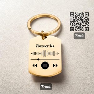 Op de afbeelding: Goudkleurige metalen sleutelhanger met een rechthoekig label. Het label heeft de tekst "Forever Us" boven een geluidsgolfafbeelding en mediabesturingspictogrammen. Een QR-code en het woord "Back" staan op de achterkant.