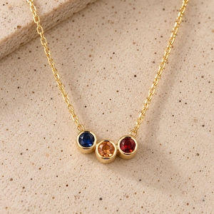 Può includere: Una collana a catena color oro con tre pietre circolari incastonate a lunetta disposte orizzontalmente. Le pietre sono di colore blu intenso, arancione e rosso scuro, ciascuna racchiusa in una montatura in metallo color oro lucido.