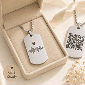 May include: Two silver-toned metal dog tag pendants on chains. One pendant features an engraved heart and a sound wave pattern. The other pendant displays a square QR code. The items are presented in a beige jewellery box with a ribbon.
