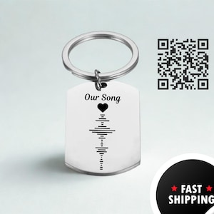 Könnte beinhalten: Ein silberfarbener Metallschlüsselanhänger mit einem rechteckigen Anhänger. Der Anhänger zeigt den Text "Our Song" über einem Herz und einer Schallwellengrafik. Ein QR-Code ist im Hintergrund sichtbar.