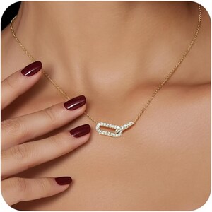 May include: A gold-toned necklace with a linked rectangular pendant encrusted with small, clear gemstones. The necklace is delicate and sits on a person's skin. The person's hand with dark red nail polish is visible.