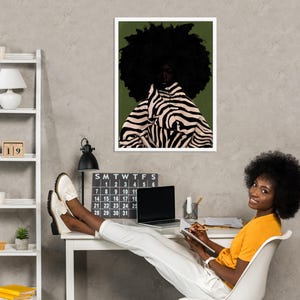 Op de afbeelding: Een ingelijste kunstdruk met een portret van een persoon met een grote afro en kleding met zebraprint. De print hangt boven een bureau met een kalender, een laptop en een persoon die op een stoel zit.