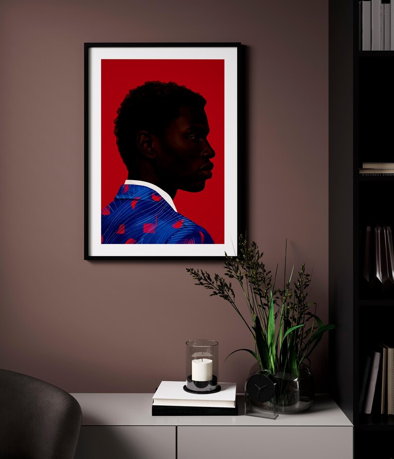 May include: Framed art print featuring a profile of a person with dark skin against a red background. The person wears a blue jacket with red accents. The artwork is displayed on a brown wall, with a candle and plant in front.