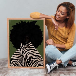 Op de afbeelding: Een ingelijste kunstdruk met een portret van een persoon met een grote, donkere afro. De persoon draagt een zwart-wit gestreept zebra-kledingstuk tegen een groene achtergrond. De lijst is lichtbruin.